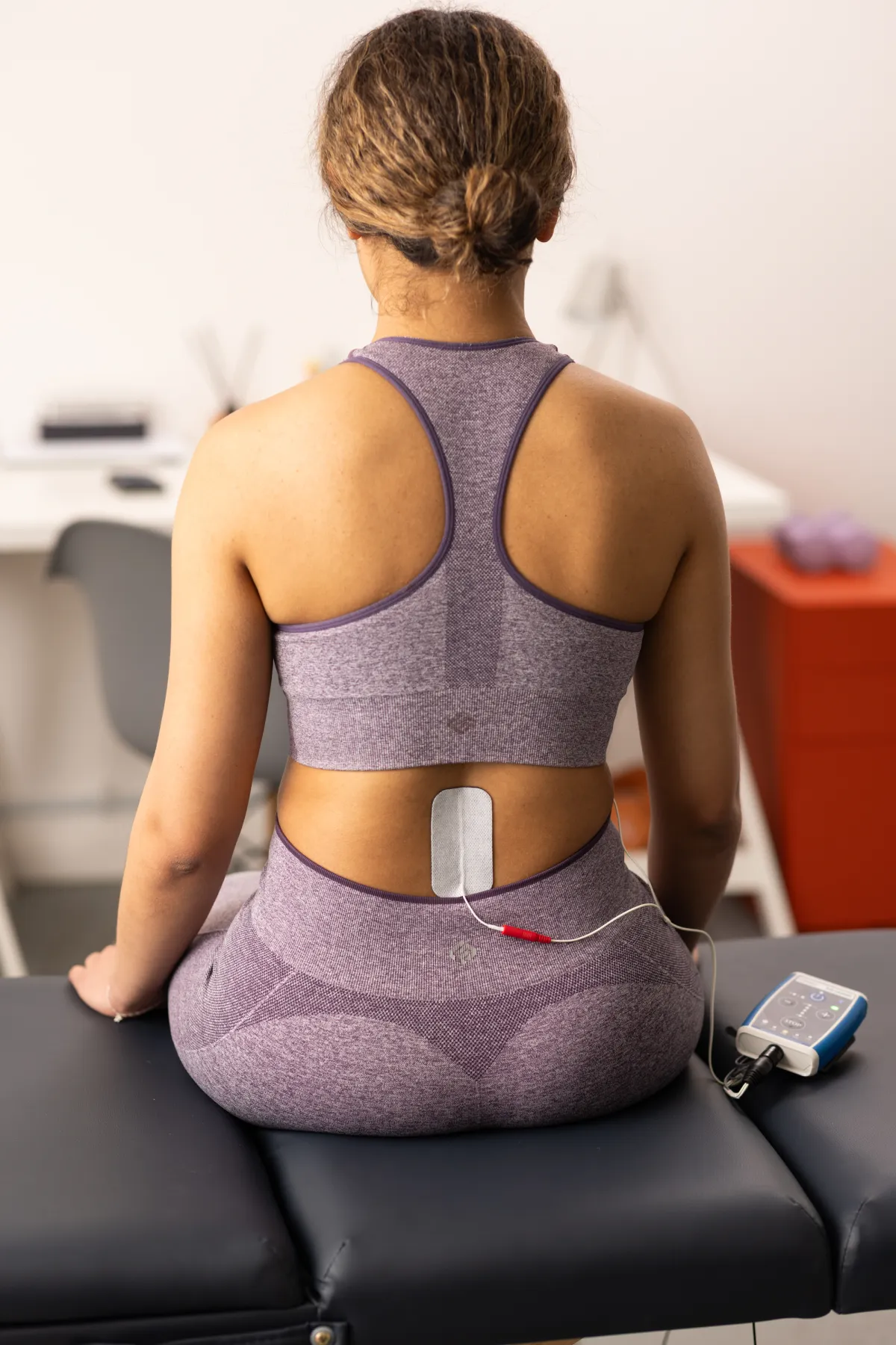 A woman seated on a treatment couch viewed from behind, with a large self-adhesive electrode over the lower back at T11/T12 connected by leads to a Stim2Go unit, set up for tSCS priming.